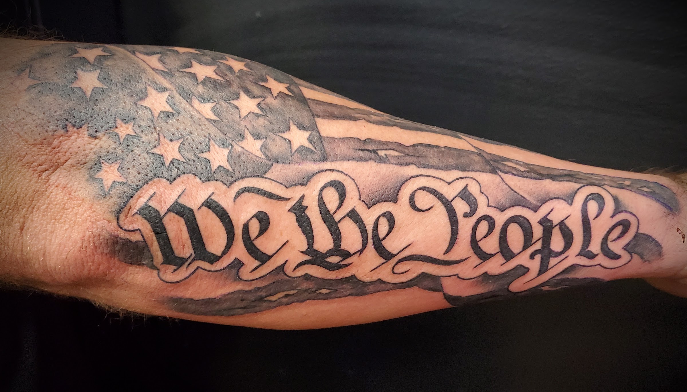 Black-and-grey We the People American flag lettering forearm tattoo